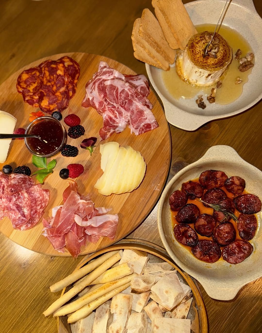 Mesa de madeira com uma tábua de enchidos, presunto e fruta, um prato de rodelas de enchido, palitos de pão e um prato com um queijo redondo, mel, nozes e pão torrado