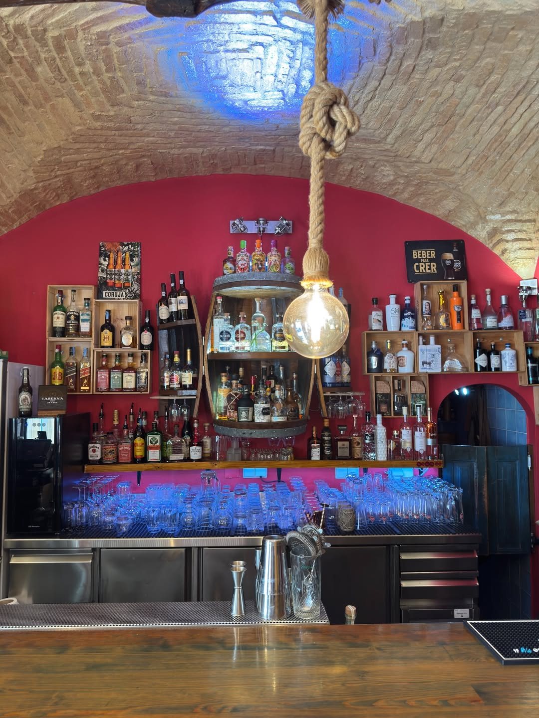 Balcão de bar com parede vermelha, prateleiras de garrafas, copos ao fundo, uma lâmpada pendurada de uma corda e teto abobadado de tijolo