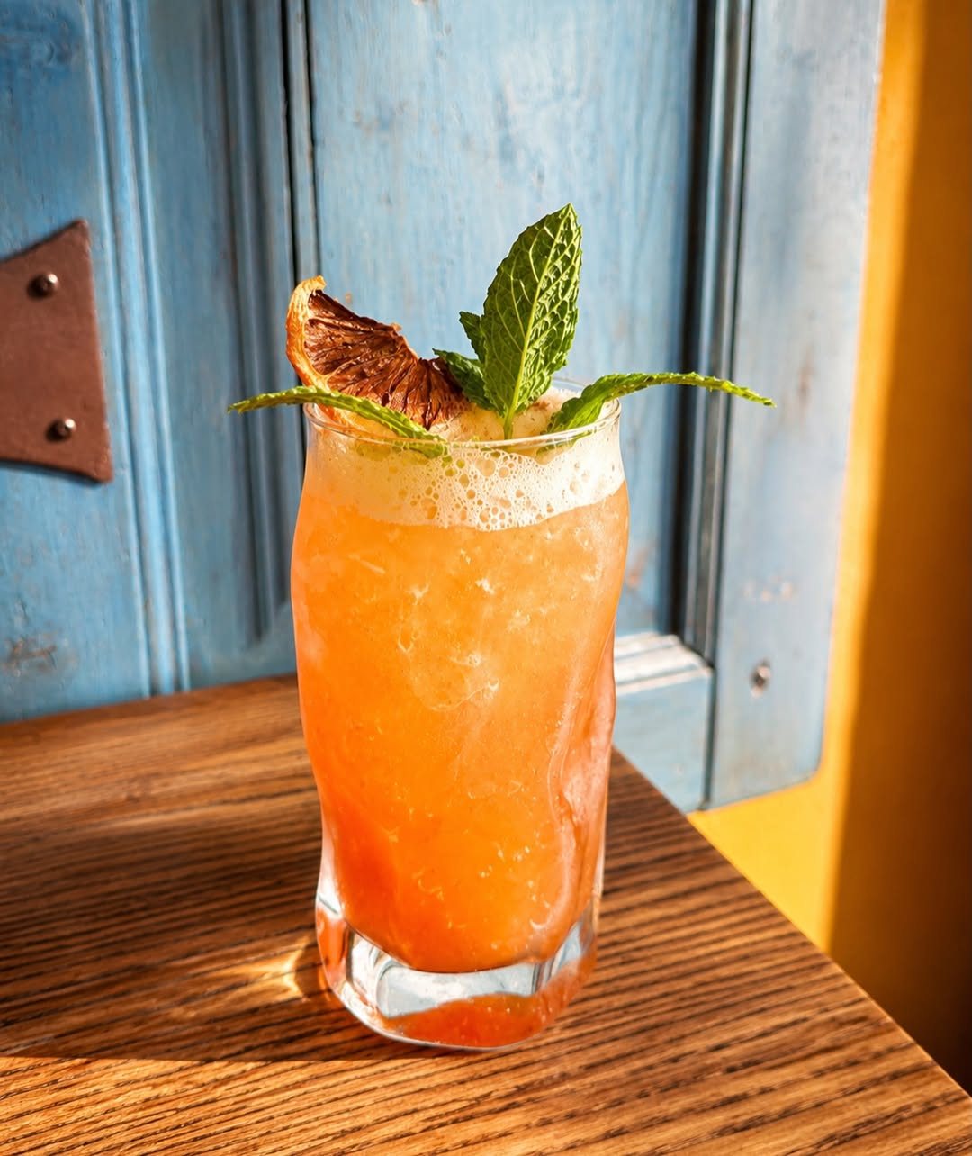 Copo com um cocktail cor de laranja, folhas de hortelã e uma fatia de fruta seca, sobre uma mesa de madeira, junto a uma porta azul
